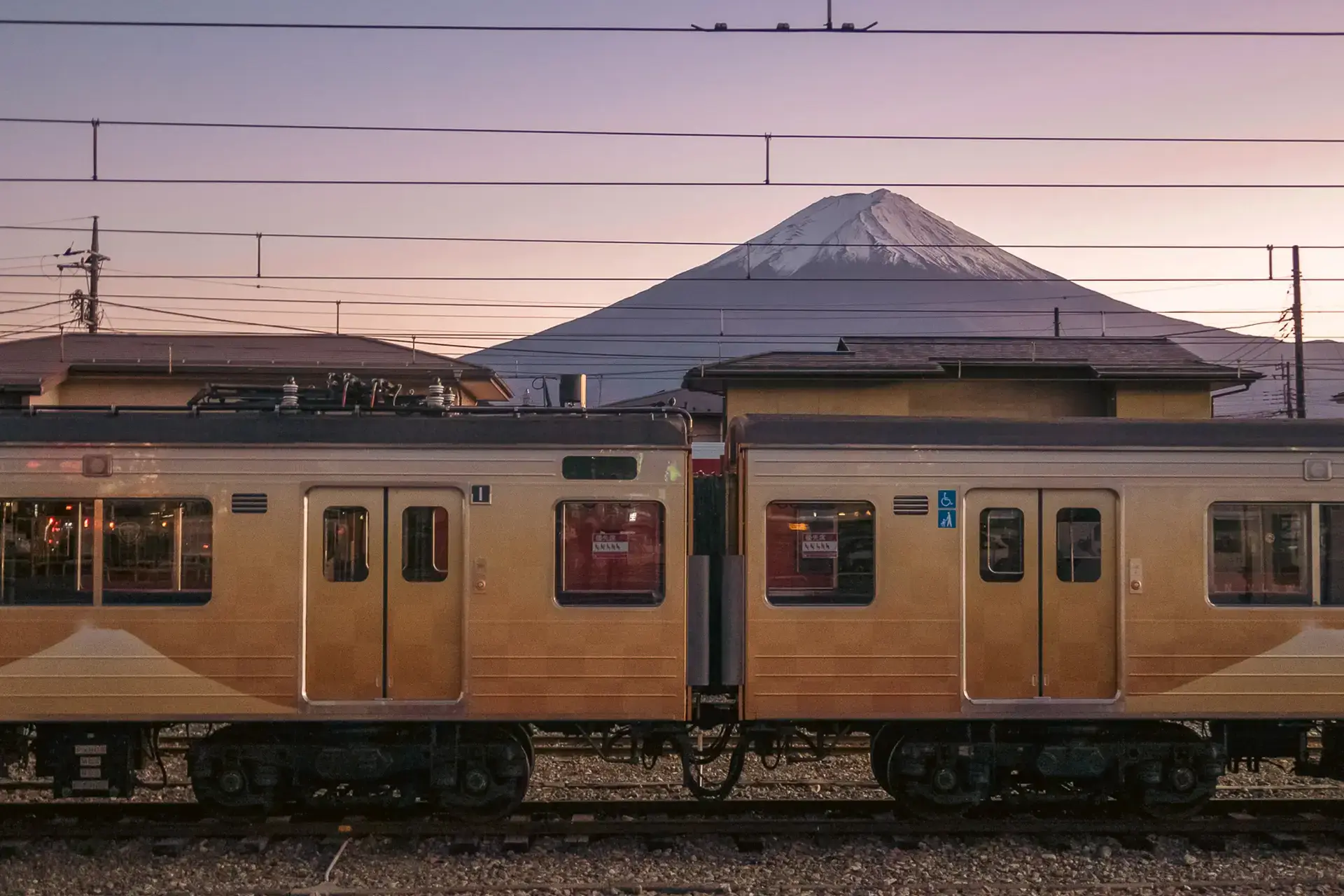 Commuter train passing through a Japanese railway station at dusk with Mount Fuji silhouetted in the background against a pink and purple sky.
