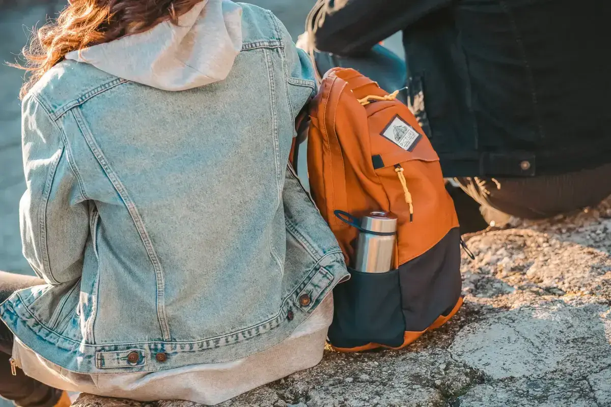 Traveler wearing denim jacket carrying orange backpack and reusable water bottle, promoting eco-friendly sustainable business travel practices.