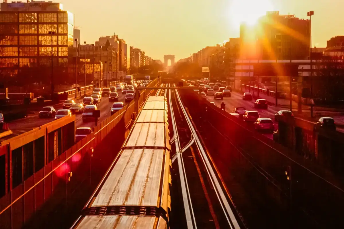 Metro train traveling through urban Paris at sunset with Arc de Triomphe visible in the distance, surrounded by city traffic and buildings bathed in golden light.