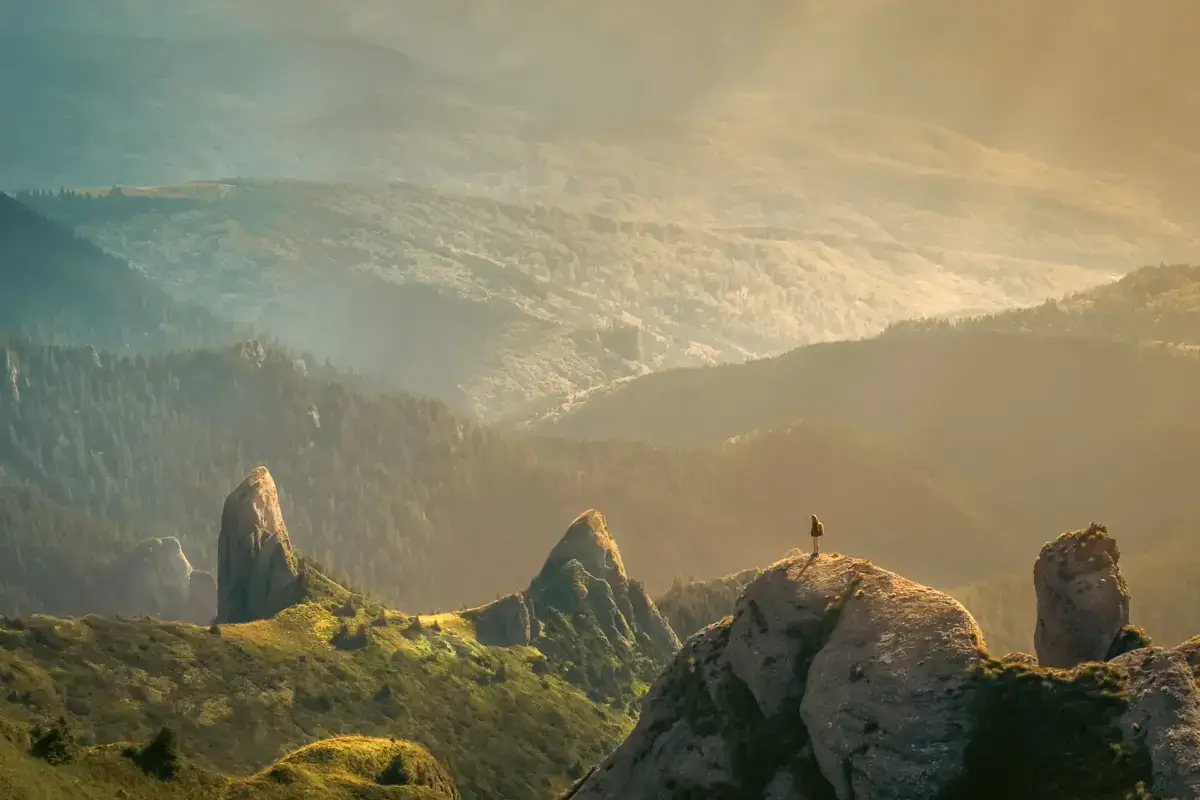 Lone hiker standing on a rocky mountain summit overlooking a vast valley with rolling hills and forests bathed in golden sunlight.