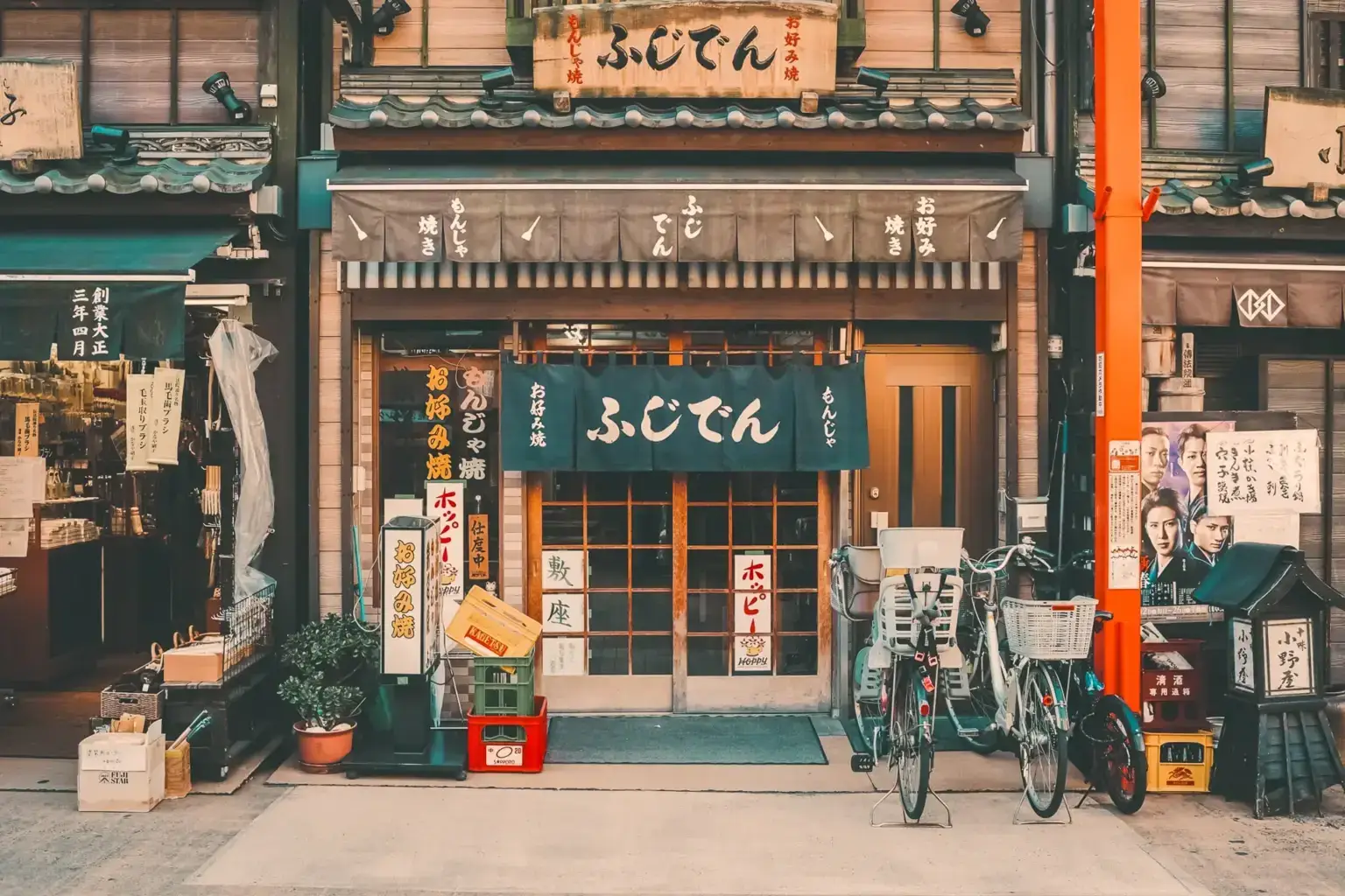 Traditional Japanese udon restaurant storefront with wooden architecture, noren curtains displaying "ふじてん" signage, and bicycles parked outside. A quintessential local neighborhood eatery showcasing authentic Japanese street culture and sustainable community-based dining.