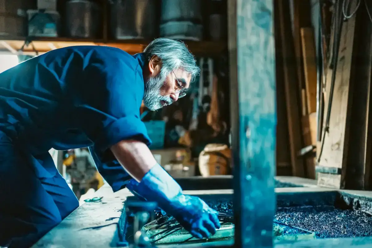 Mature craftsman with gray beard carefully works on metal piece in traditional workshop, surrounded by tools and bathed in natural light.