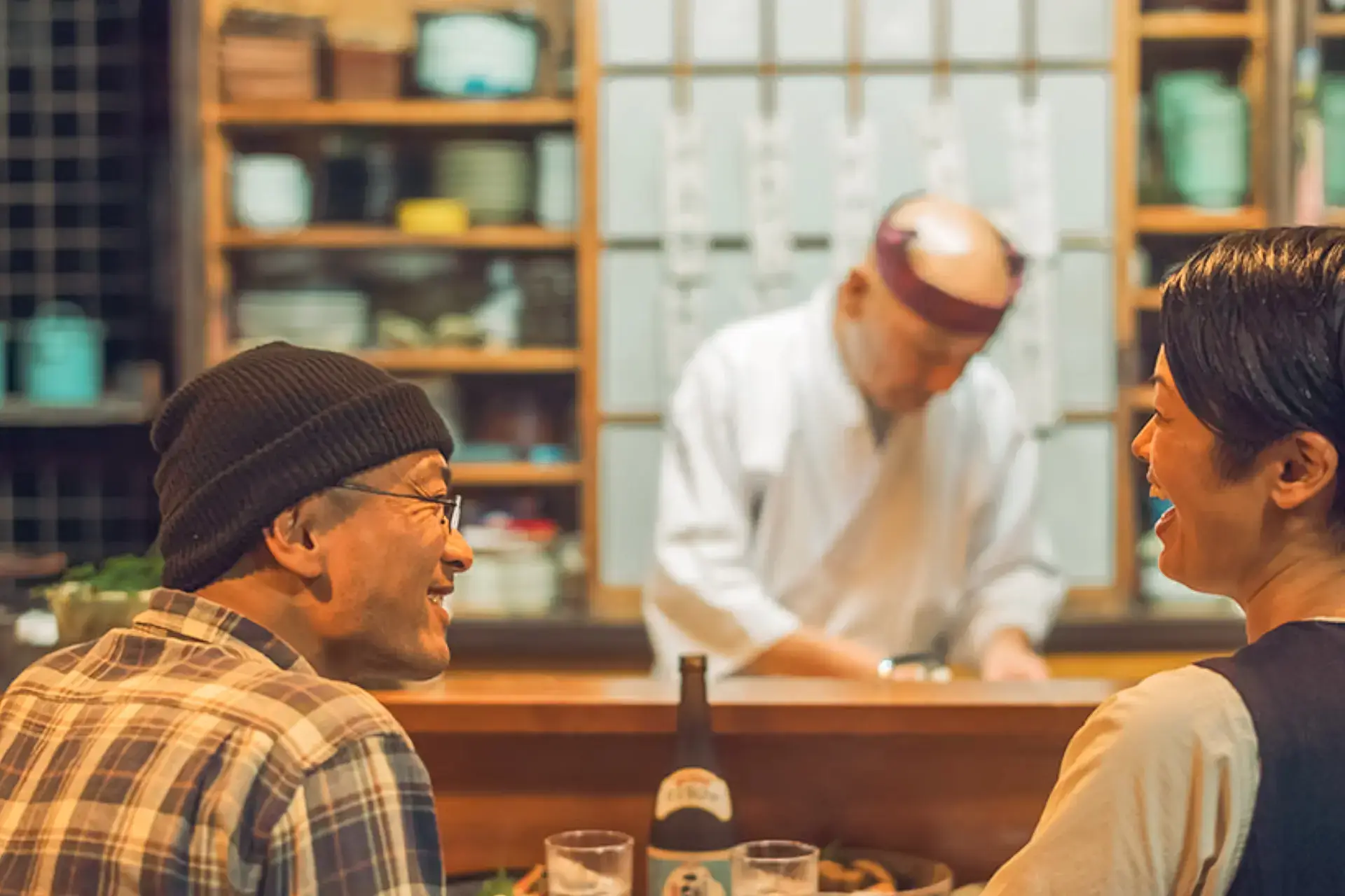 Two customers enjoying conversation at a traditional Japanese restaurant counter while a chef prepares food in the background.