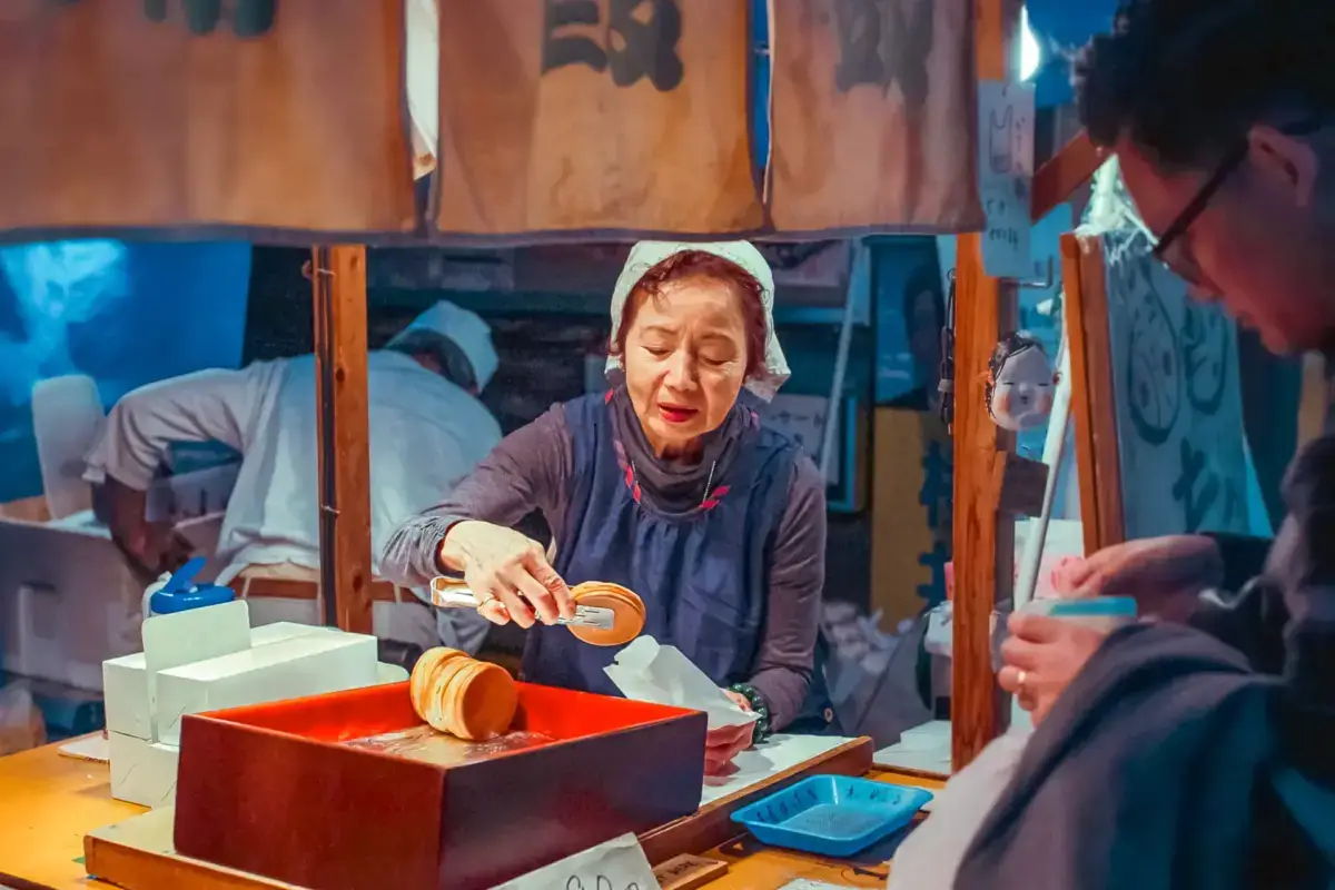 Japanese woman in traditional white headscarf demonstrating rice cake making at a local Yatsugatake tourism event, using traditional wooden tools with customers watching nearby.