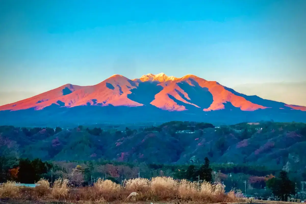 Yatsugatake mountain range glowing with orange and pink alpenglow at sunrise or sunset, viewed over forested hills and residential areas below a clear blue sky.