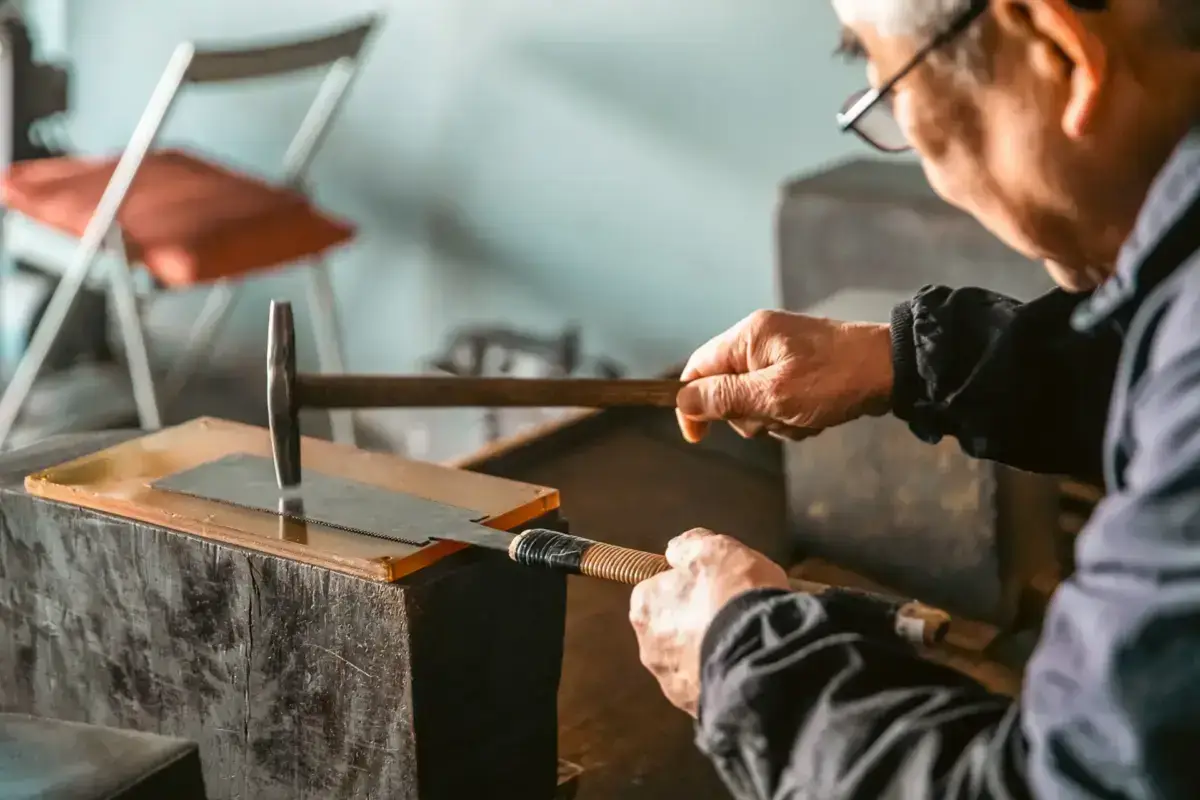 Elderly craftsman using traditional hand saw on wooden board mounted on rustic workbench in Japanese workshop, demonstrating traditional woodworking techniques supporting local artisan economy in Yatsugatake.