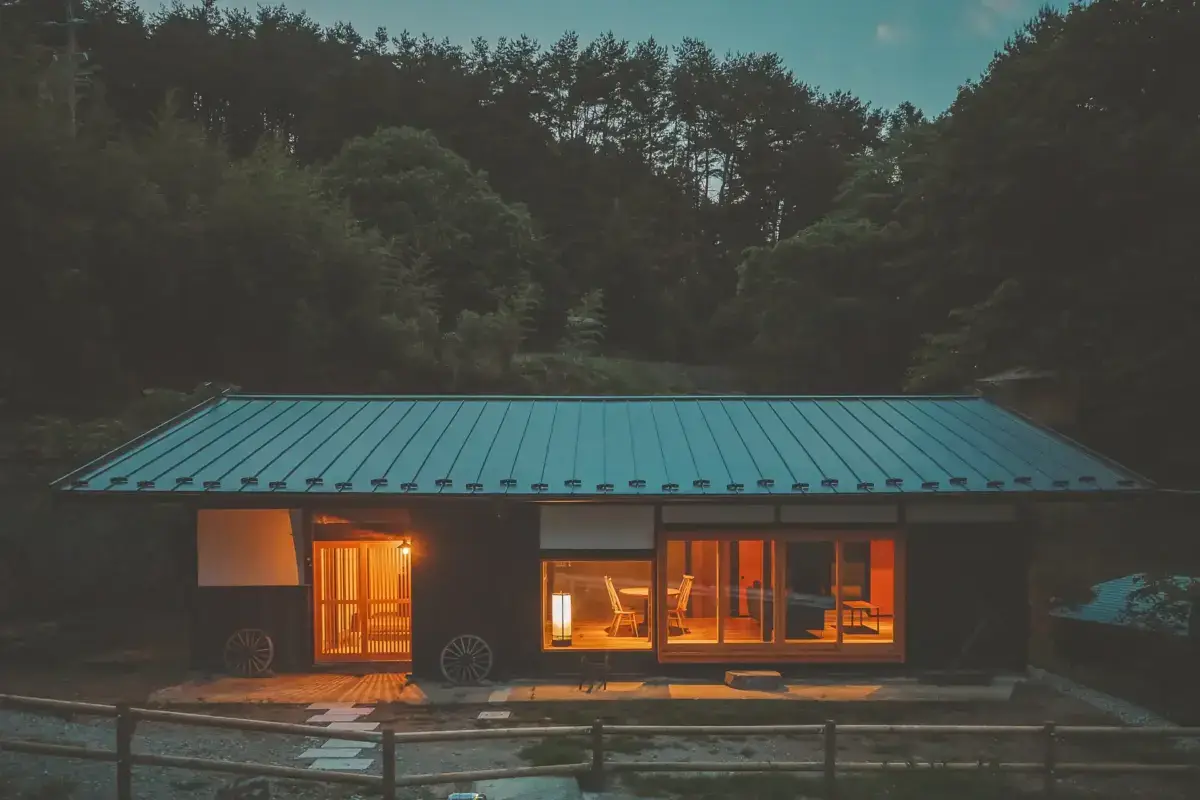 Modern Japanese-style vacation cottage with blue metal roof and warmly lit interior at dusk, nestled in forested Yatsugatake mountains with traditional sliding doors and wooden deck.