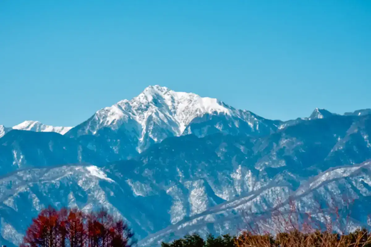 Snow-capped mountain peaks rising above forested ridges under a clear blue sky, showcasing natural Japanese alpine landscape ideal for sustainable tourism and community development.
