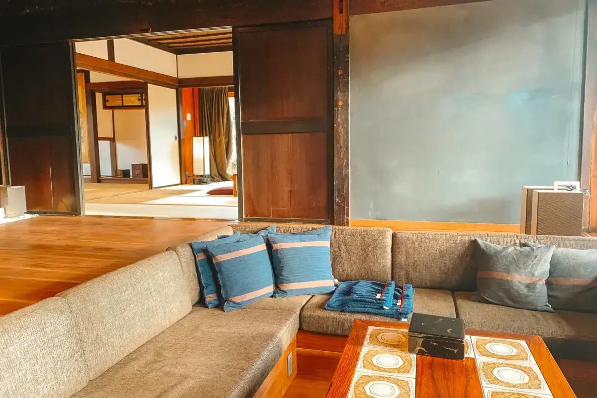Modern Japanese interior lounge area with beige sectional sofa featuring blue and gray cushions, wooden coffee table, and traditional tatami room visible through sliding doors in the background.