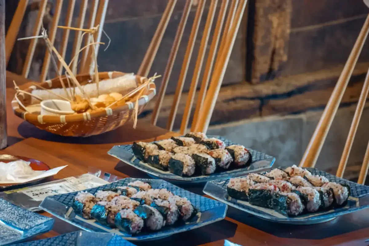 Traditional Japanese onigiri rice balls with nori seaweed arranged on blue ceramic plates, served alongside a bamboo basket of corn at an outdoor cafe with wooden chairs.