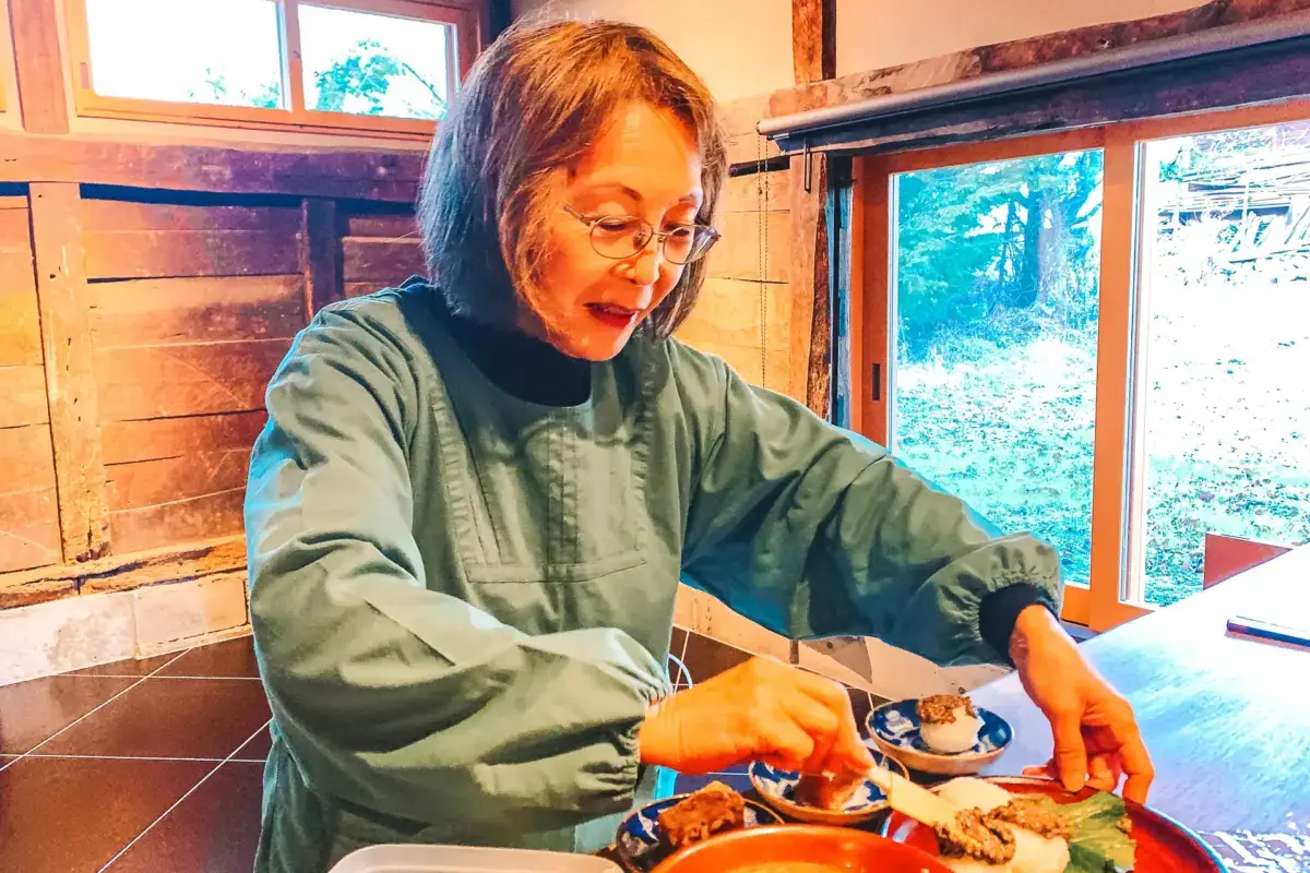 Woman wearing glasses and green traditional clothing enjoying a Japanese meal served on lacquerware trays in a rustic wooden building with natural forest views through the windows.