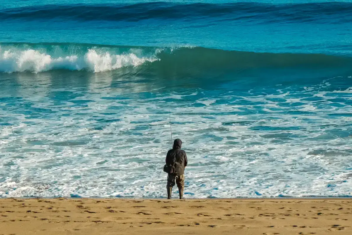 Solitary fisherman standing on sandy beach casting into turquoise ocean waves, representing coastal marine resource utilization and sustainable blue economy practices.