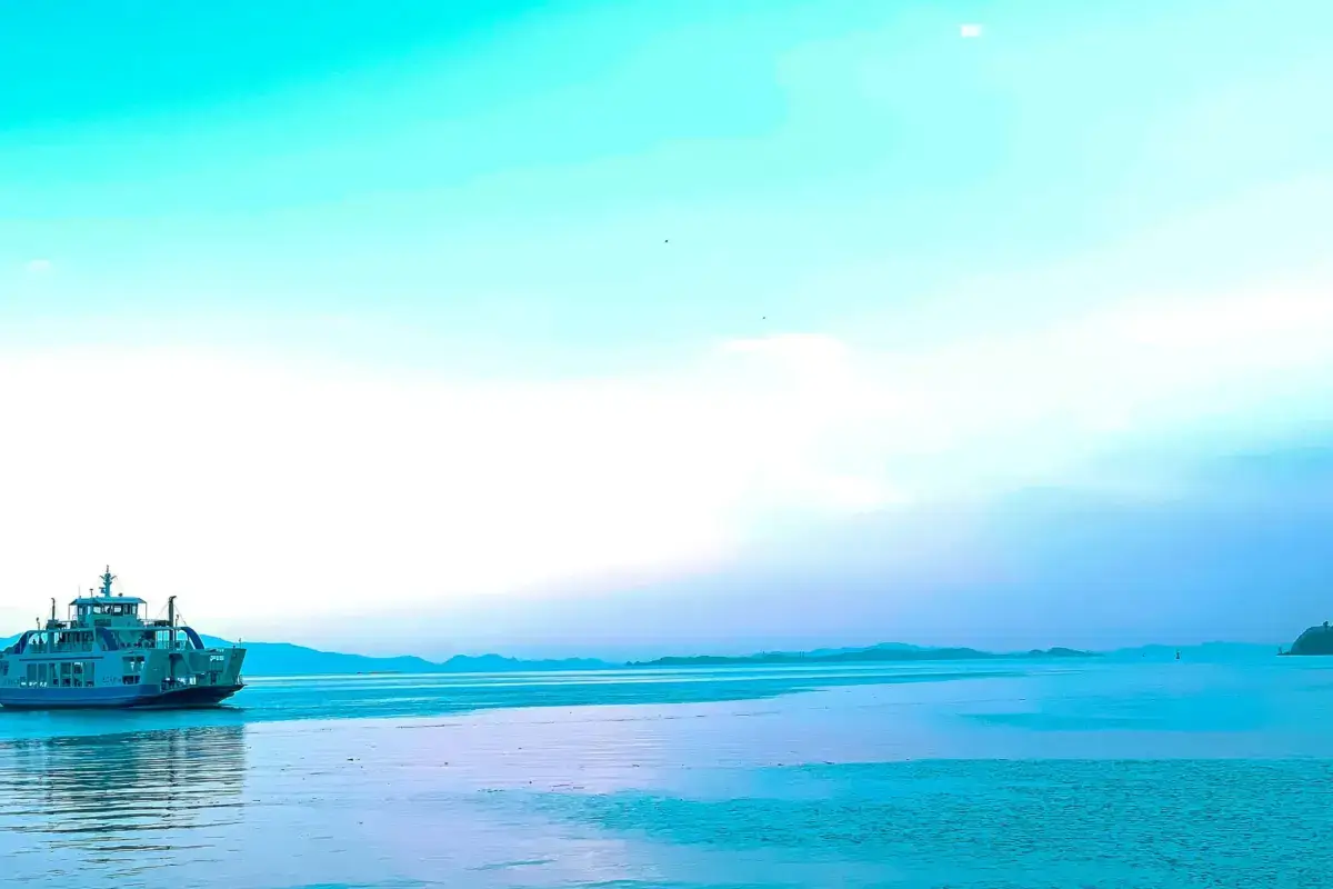 Ferry boat sailing across calm blue waters with distant mountain islands on the horizon under a clear turquoise sky in Japan