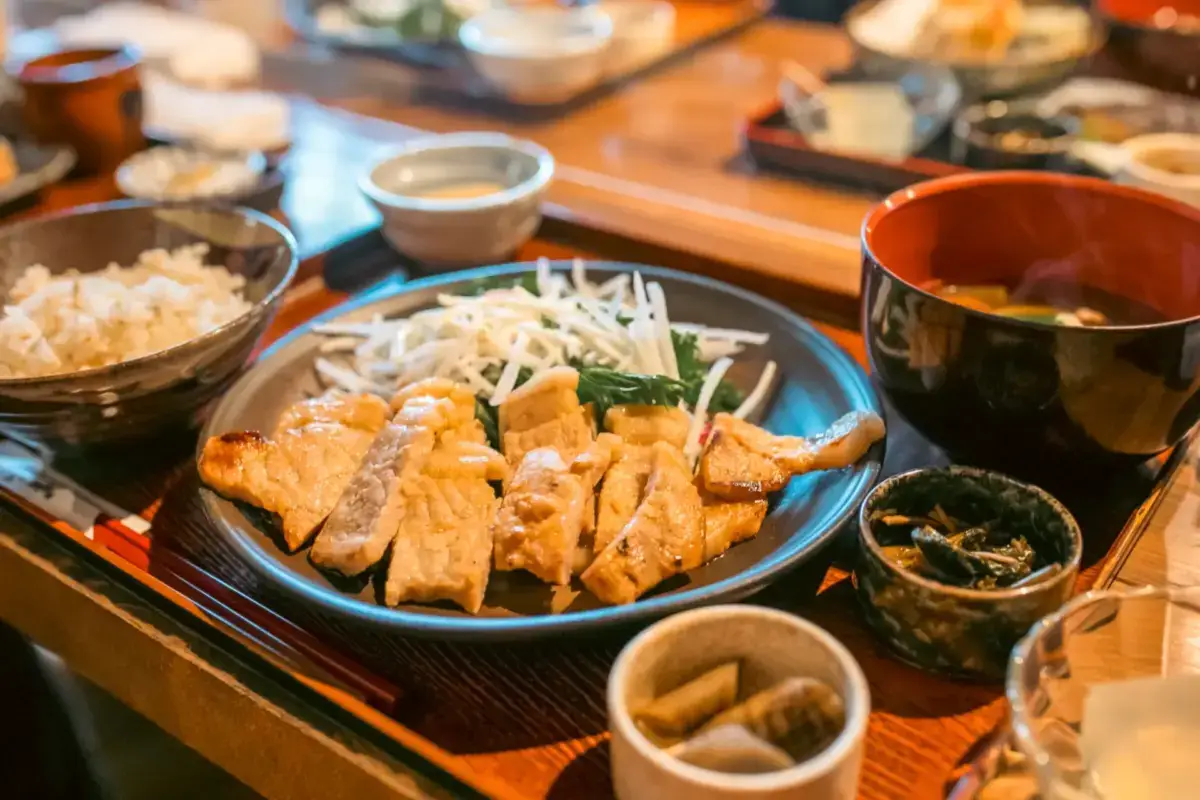 Japanese-style meal set featuring grilled chicken or tofu with bean sprouts and vegetables on a blue tray, accompanied by steamed rice, miso soup, and assorted side dishes.