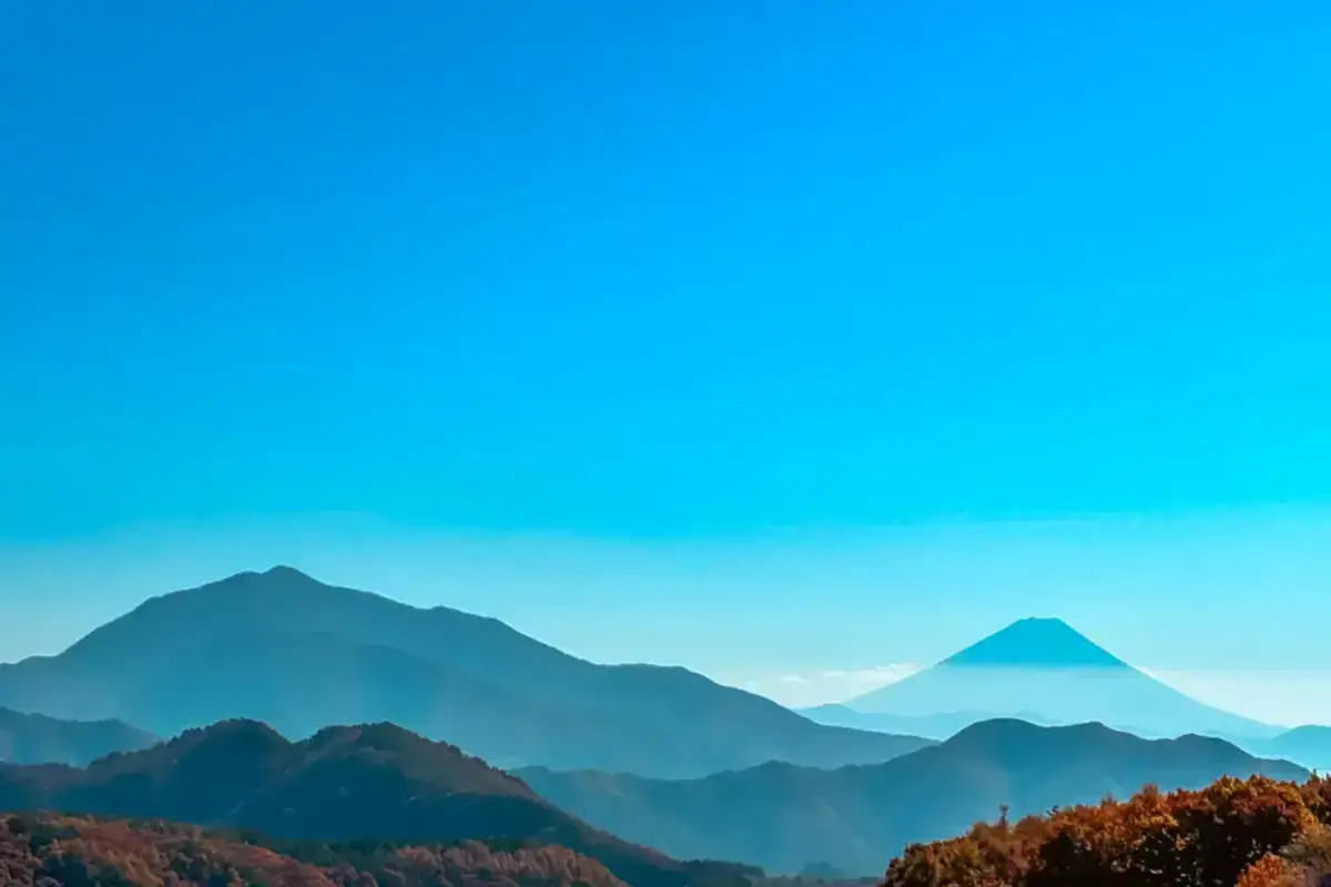 Layered mountain ranges with autumn foliage in the foreground beneath a bright blue sky, featuring a distinctive conical peak in the distance.