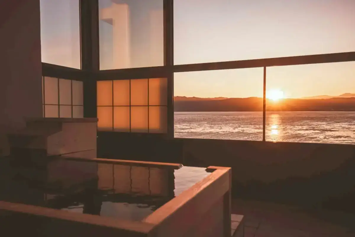 Tranquil indoor hot tub with large windows overlooking ocean sunset, featuring traditional Japanese-style sliding doors and warm ambient lighting creating a luxurious spa atmosphere.