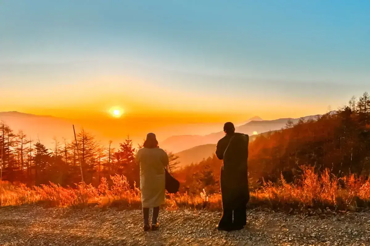 Two people with backpacks watching a golden sunrise over misty mountain ranges from a scenic overlook, surrounded by silhouetted pine trees and autumn grasses.