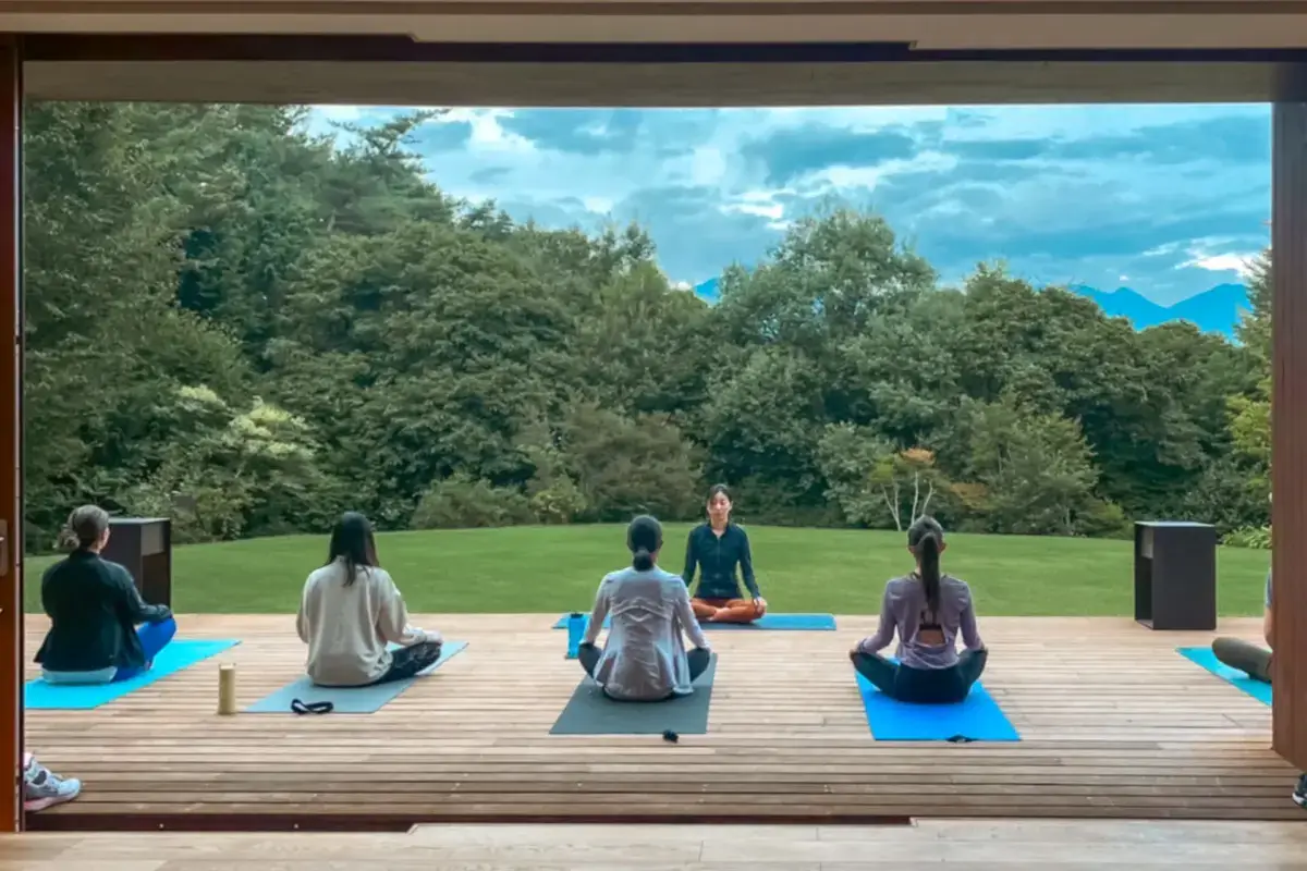 Outdoor yoga class with instructor leading four participants in meditation pose on blue mats on a wooden deck, surrounded by lush green trees and mountain views under a bright blue sky.