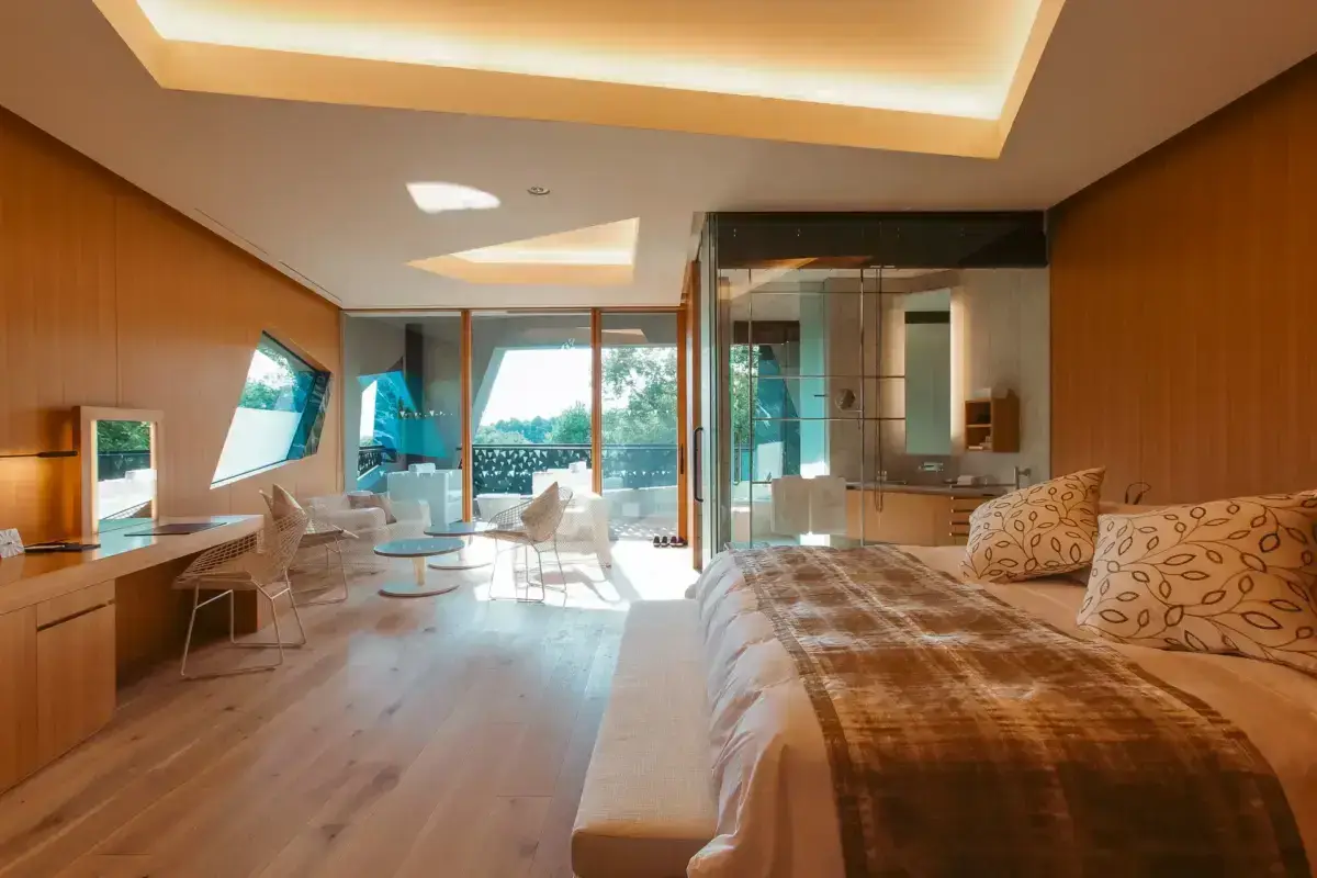 Luxurious modern hotel suite with floor-to-ceiling windows opening to a private terrace, featuring warm wood interiors, a comfortable bed with decorative pillows, and a glass-enclosed bathroom with ambient lighting.
