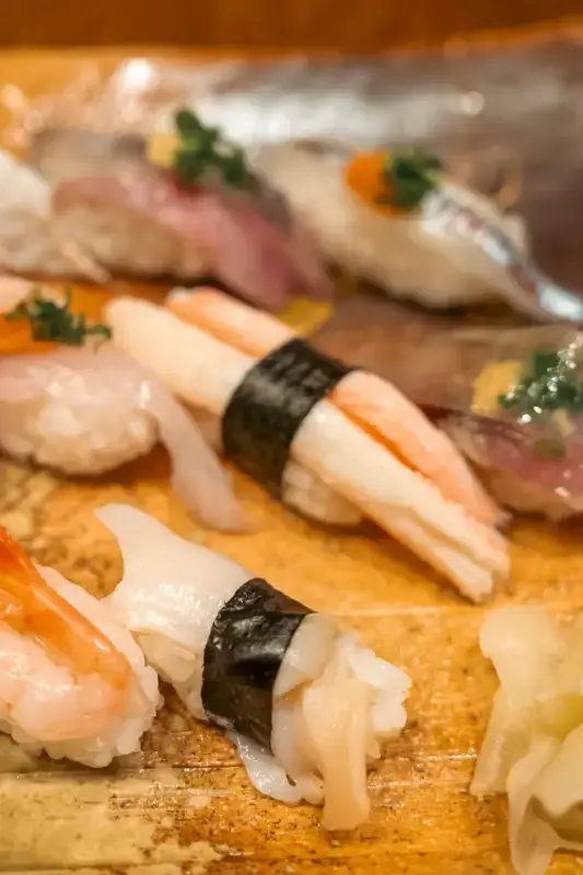Assorted nigiri sushi arranged on a wooden board, featuring various fresh fish and seafood topped with garnishes, including tuna, salmon, and squid wrapped with nori seaweed.