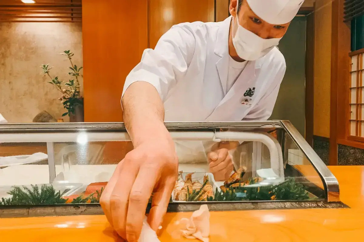 Japanese sushi chef in white uniform and face mask carefully preparing fresh fish at a wooden counter in front of a refrigerated display case of seafood