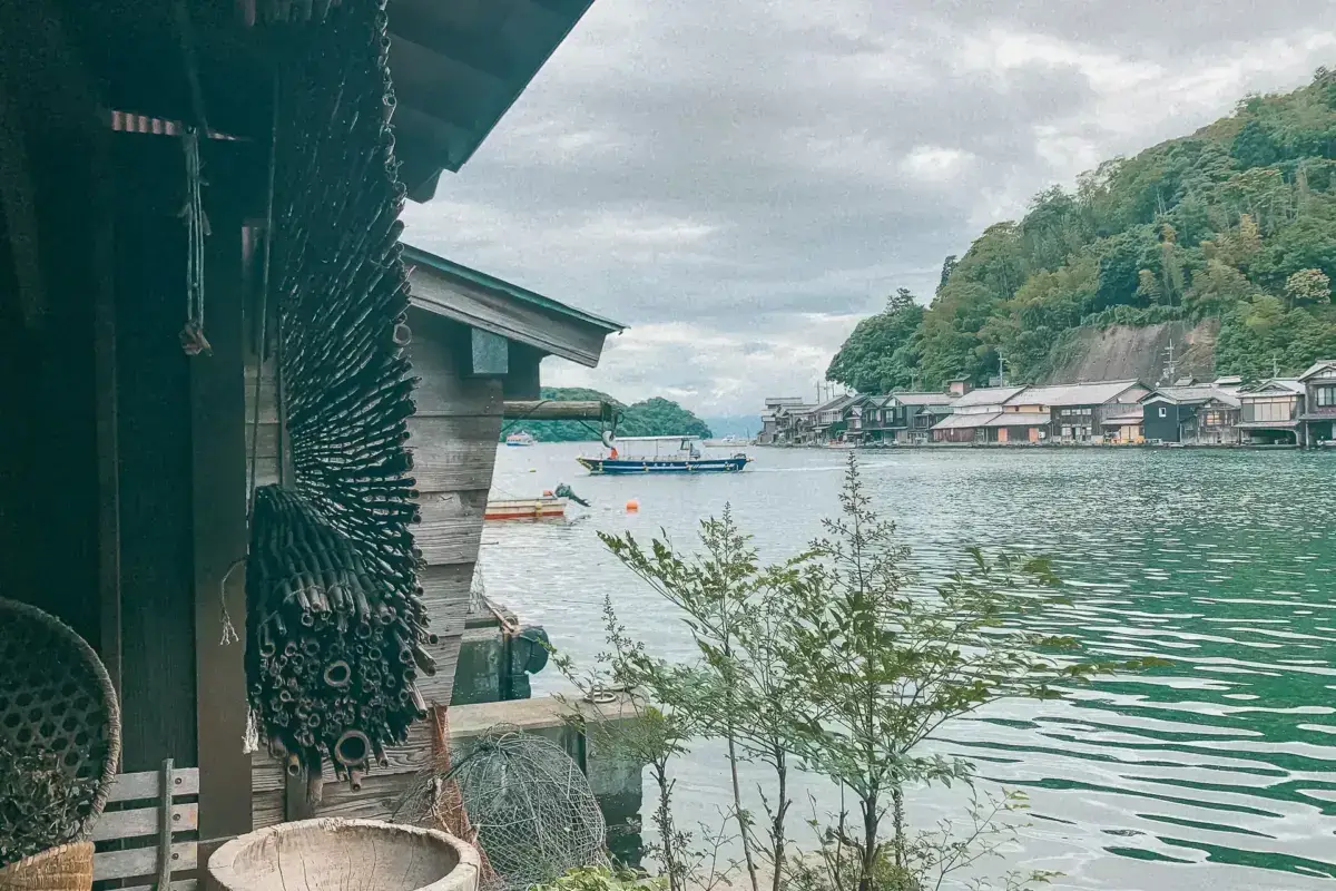 Traditional Japanese coastal fishing village with wooden boathouses (funaya) built over calm green waters, showing a small fishing boat moored in the bay surrounded by forested hills under cloudy skies.