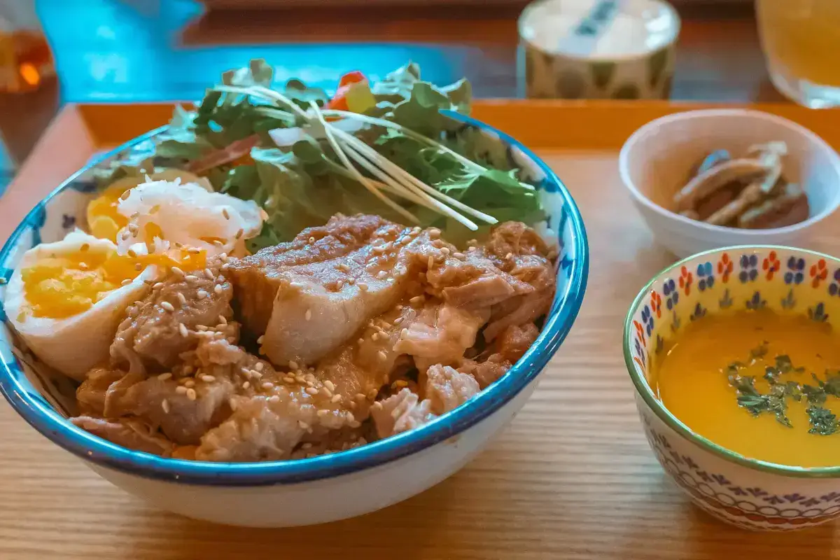 Japanese pork and rice bowl with soft-boiled egg, fresh greens, and sesame seeds served with miso soup and pickled vegetables.