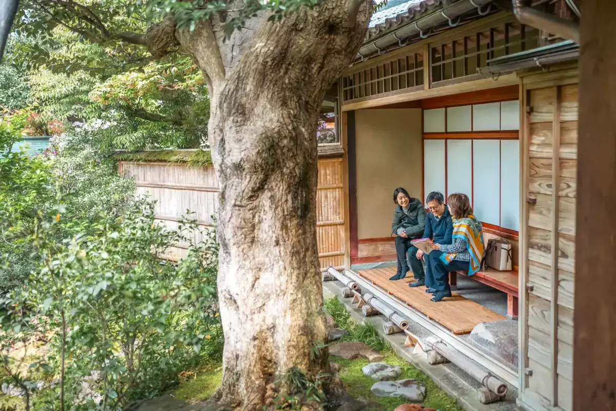 Three people sitting on the engawa porch of a traditional Japanese house with shoji screens, beside a large tree in a lush garden.