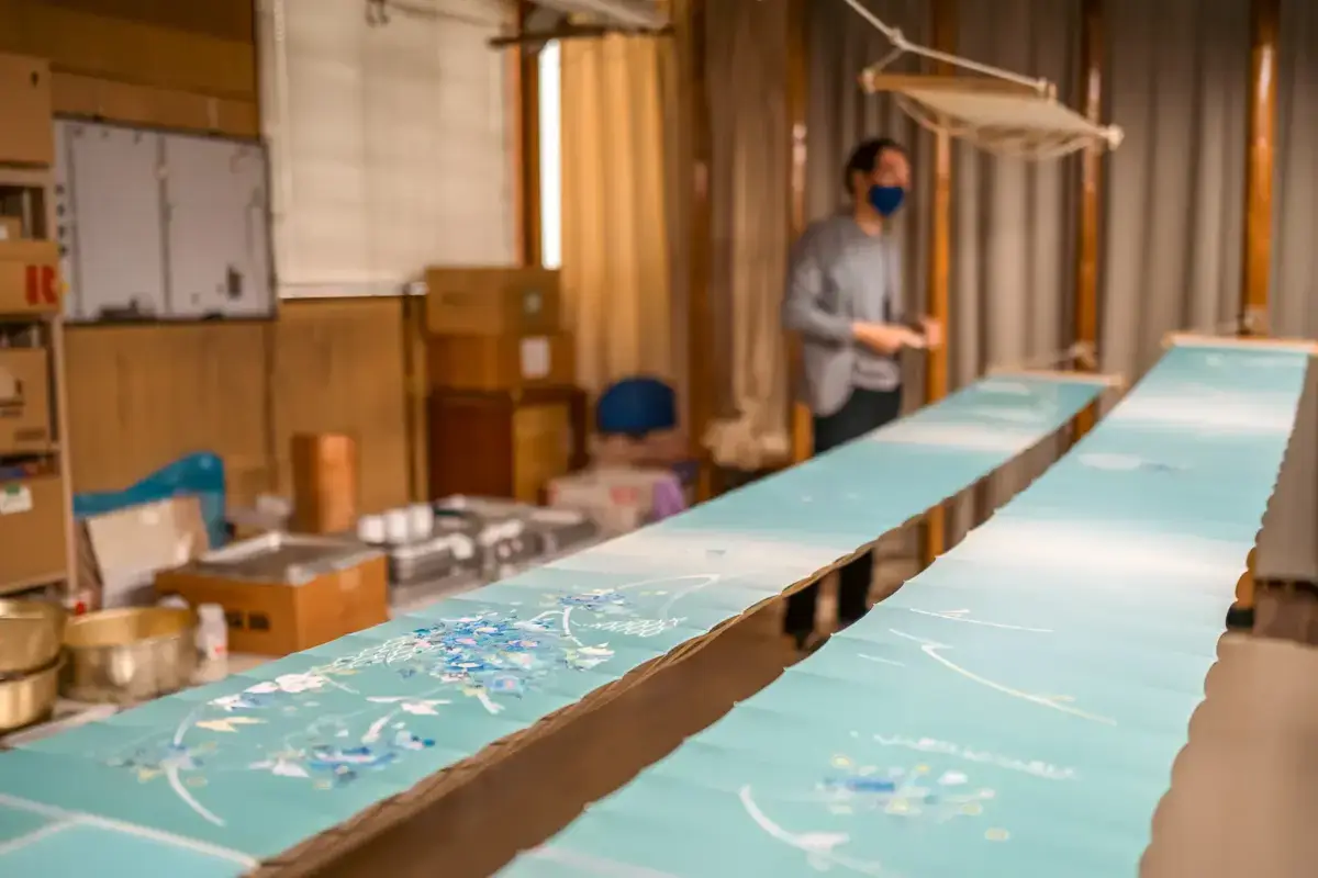 Artist in workshop painting turquoise panels with delicate white and blue floral patterns, traditional Japanese craftsmanship in progress.