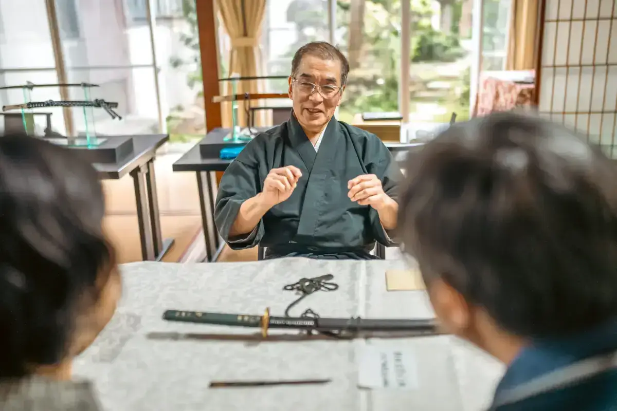 Elderly Japanese craftsman in traditional dark kimono speaking to visitors across a table displaying decorative swords and traditional metalwork in a bright, traditional Japanese room with shoji screens and garden views.