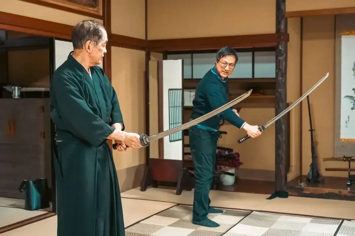 Two practitioners in traditional Japanese clothing demonstrate samurai sword techniques in a traditional tatami room with shoji screens.
