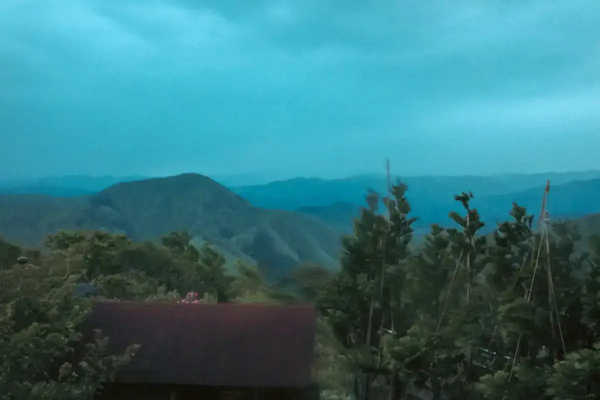 Misty mountain landscape viewed from a train window in Ishikawa, with layered mountain ridges fading into the blue-gray distance, a traditional Japanese farmhouse roof in the foreground, and lush green vegetation.