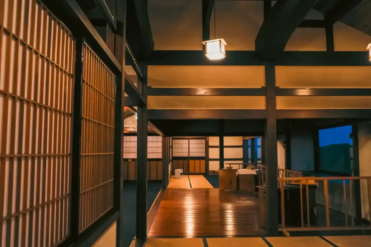 Traditional Japanese interior with exposed dark wooden beams, latticed shoji screens, warm ambient lighting, and polished wooden floors creating a serene atmospheric space.