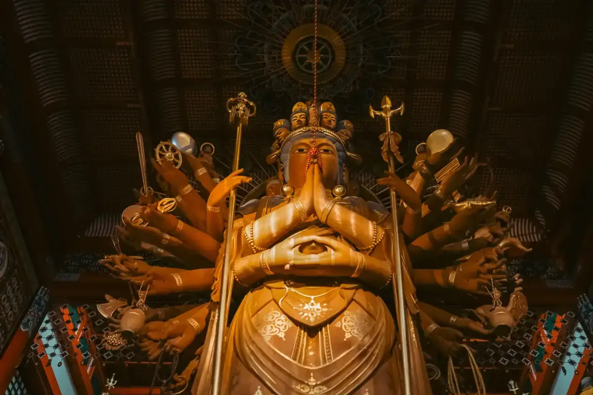 Golden multi-armed Buddhist deity statue in meditation pose with ornate decorations and symbolic objects, displayed against an intricate dark patterned backdrop with suspended ornamental disc above.