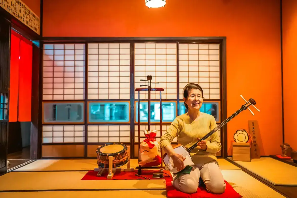 Smiling woman in yellow sweater kneeling on tatami floor while holding a shamisen, a traditional Japanese three-stringed instrument, in a room with orange walls and translucent shoji screens.