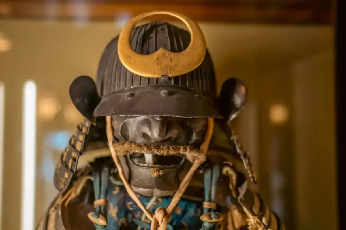 Close-up of a traditional Japanese samurai helmet and armor display featuring a black iron helmet with distinctive crescent-shaped ornament and face mask, with blue and gold-toned chest armor visible below.