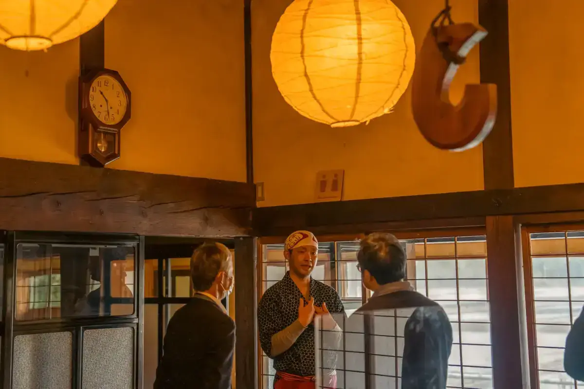 Three people conversing in a traditional Japanese interior with warm paper lanterns, exposed wooden beams, a vintage wall clock, and shoji screen doors illuminated by natural light.