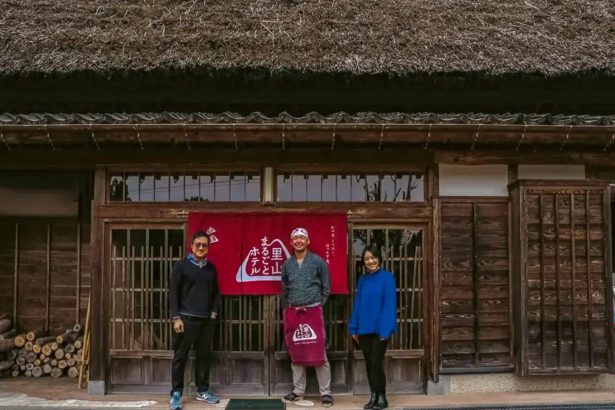 Three people standing in front of a traditional Japanese thatched-roof house with weathered wooden facade, with the center person holding a red banner with Japanese calligraphy and a pink tote bag.