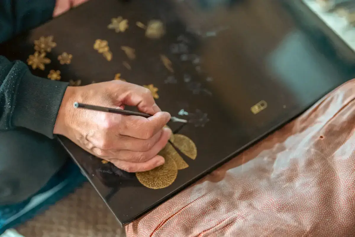 Artisan's hands carefully painting delicate gold floral motifs onto a black lacquerware tray, with a pink decorative fabric visible underneath.