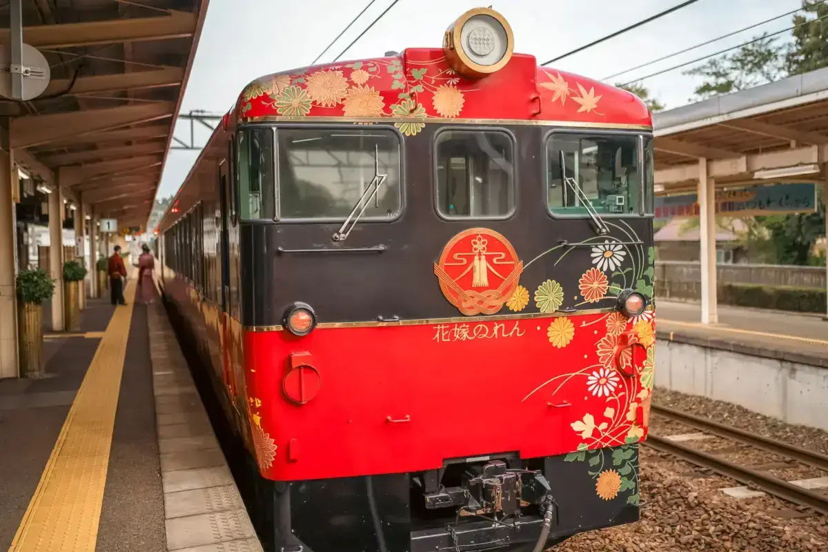 Brightly decorated Japanese sightseeing train with red exterior and gold floral patterns stationed at a platform in Ishikawa, featuring traditional chrysanthemum and maple leaf designs.