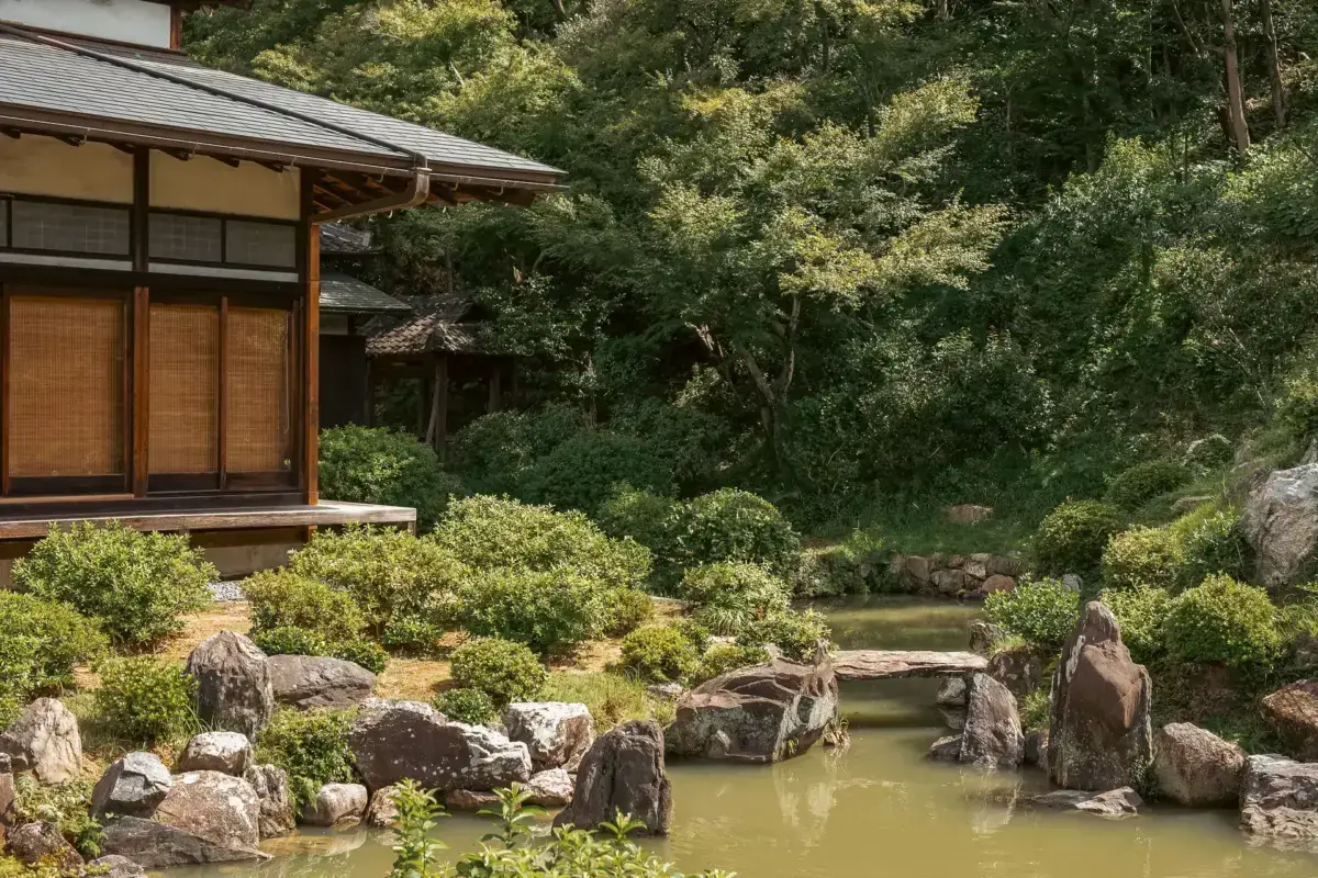 Traditional Japanese temple building with wooden architecture overlooking a serene pond garden featuring manicured shrubs, stone arrangements, and lush green forest backdrop in Yoshino.