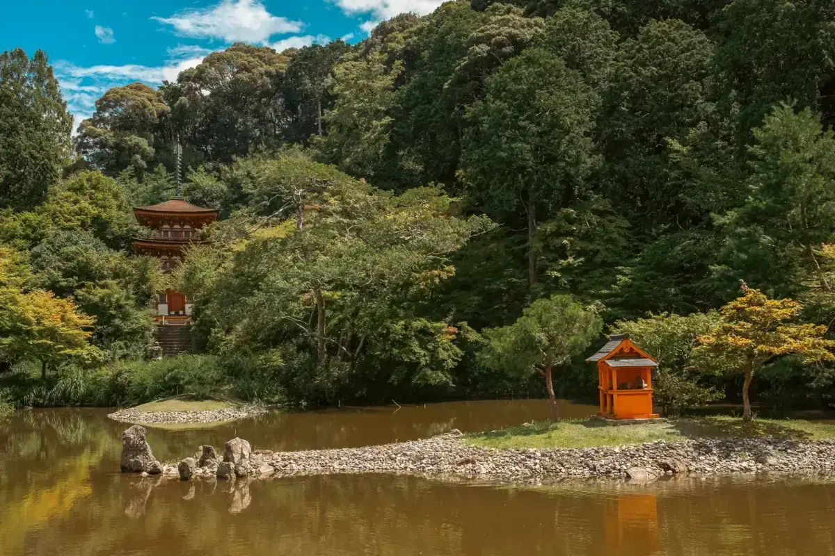 Traditional orange torii gate and red pagoda beside a tranquil pond surrounded by lush green forest in Yoshino, Japan.