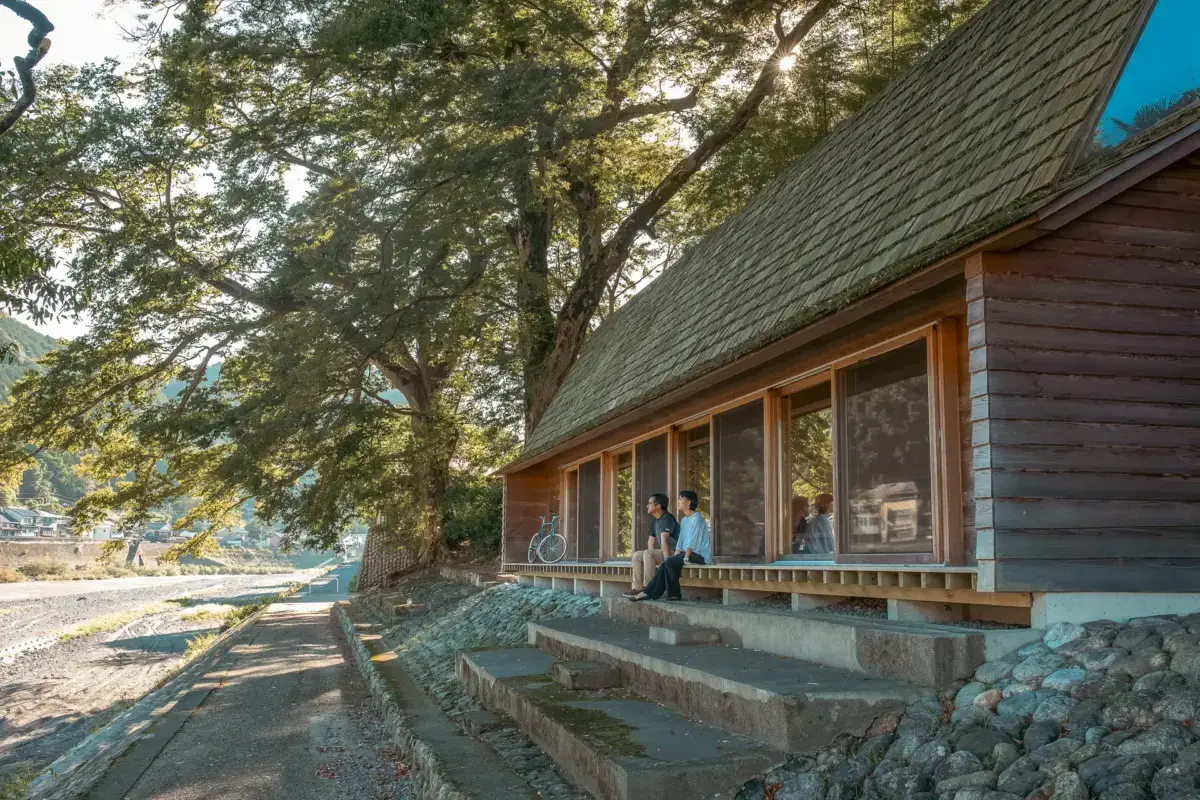 Traditional wooden Japanese building with wide veranda seating beneath a large tree, with a person sitting outside enjoying the peaceful rural setting in warm afternoon sunlight.