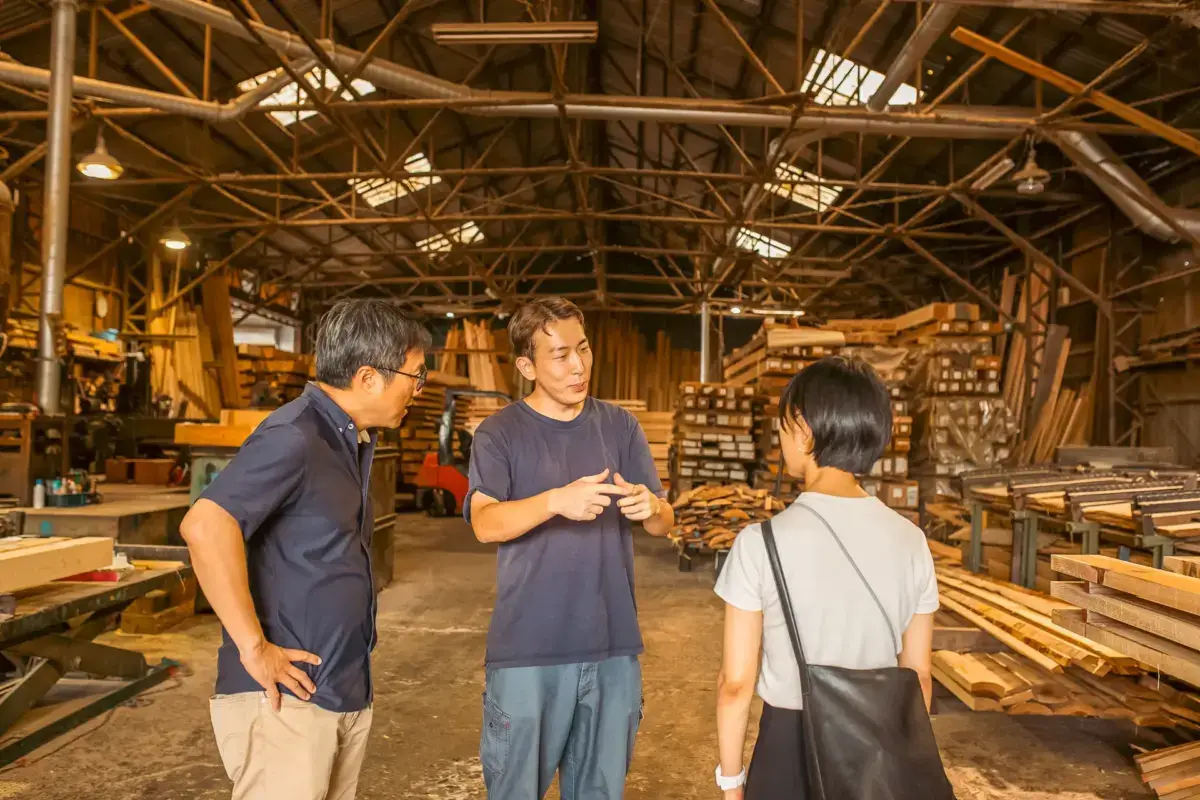 Three people conversing in a traditional Japanese woodworking workshop with exposed wooden beams, stacked lumber, and organized tools.