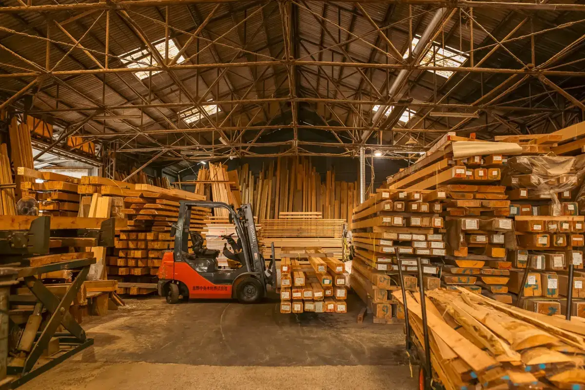 Interior of a timber warehouse with stacked lumber organized on shelves and a red forklift positioned among the wooden planks under exposed beam ceiling with industrial lighting.