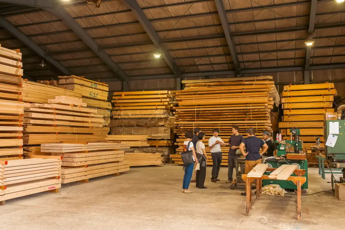 Interior of a traditional lumber warehouse in Yoshino with visitors observing stacks of milled wood planks and craftspeople working at woodworking stations.