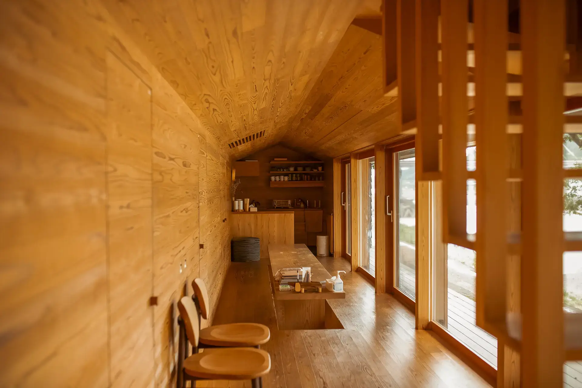 Warm plywood interior of a minimalist Japanese cabin featuring curved barrel vault ceiling with natural wood grain patterns, built-in shelving, floor-to-ceiling windows overlooking a wooden deck, and simple wooden furniture bathed in natural light.