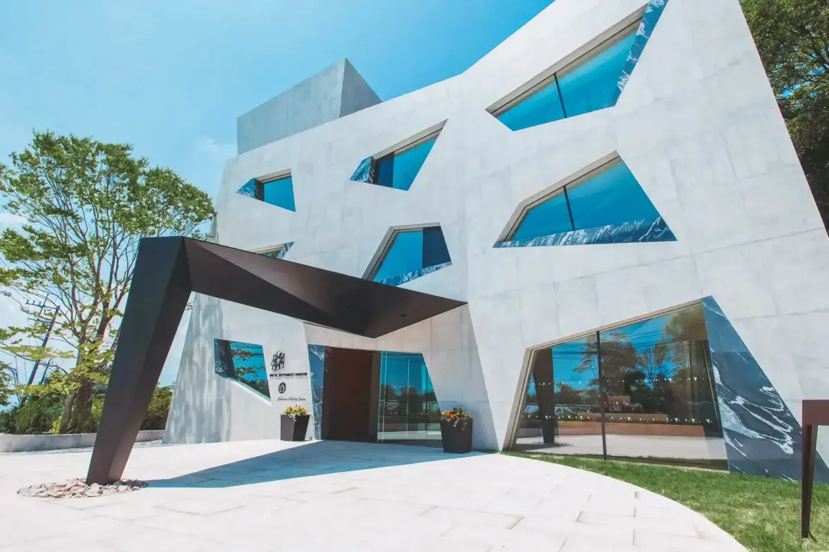 Modern white concrete building with angular geometric design featuring irregular trapezoid-shaped windows and a prominent angled metal entrance canopy, set against a clear blue sky.