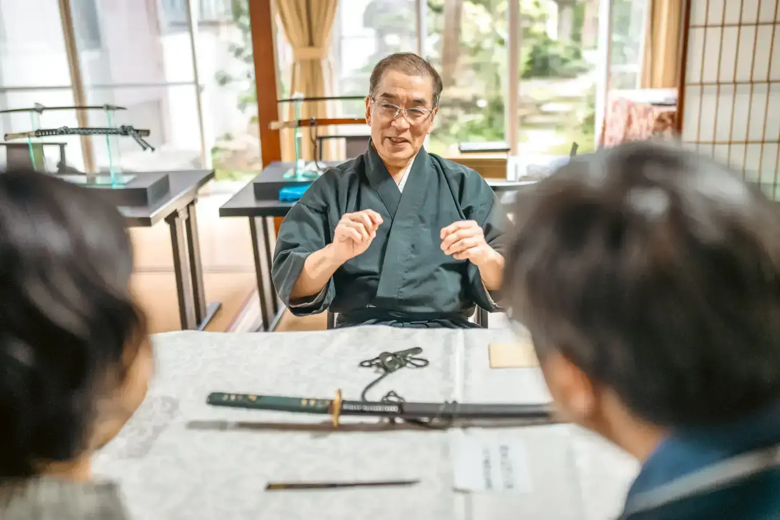 Japanese craftsman in traditional kimono demonstrating sword-making techniques to tourists at a table with katana and sword-making tools in a traditional Japanese room.