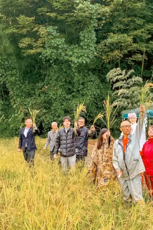 Group of people walking through golden autumn field carrying harvested rice stalks, with lush green forest in the background